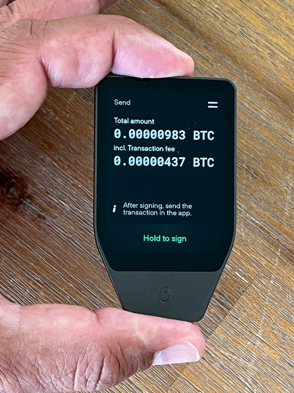 A hardware wallet held between two fingers, its screen showing a transaction summary and a Hold to sign prompt
