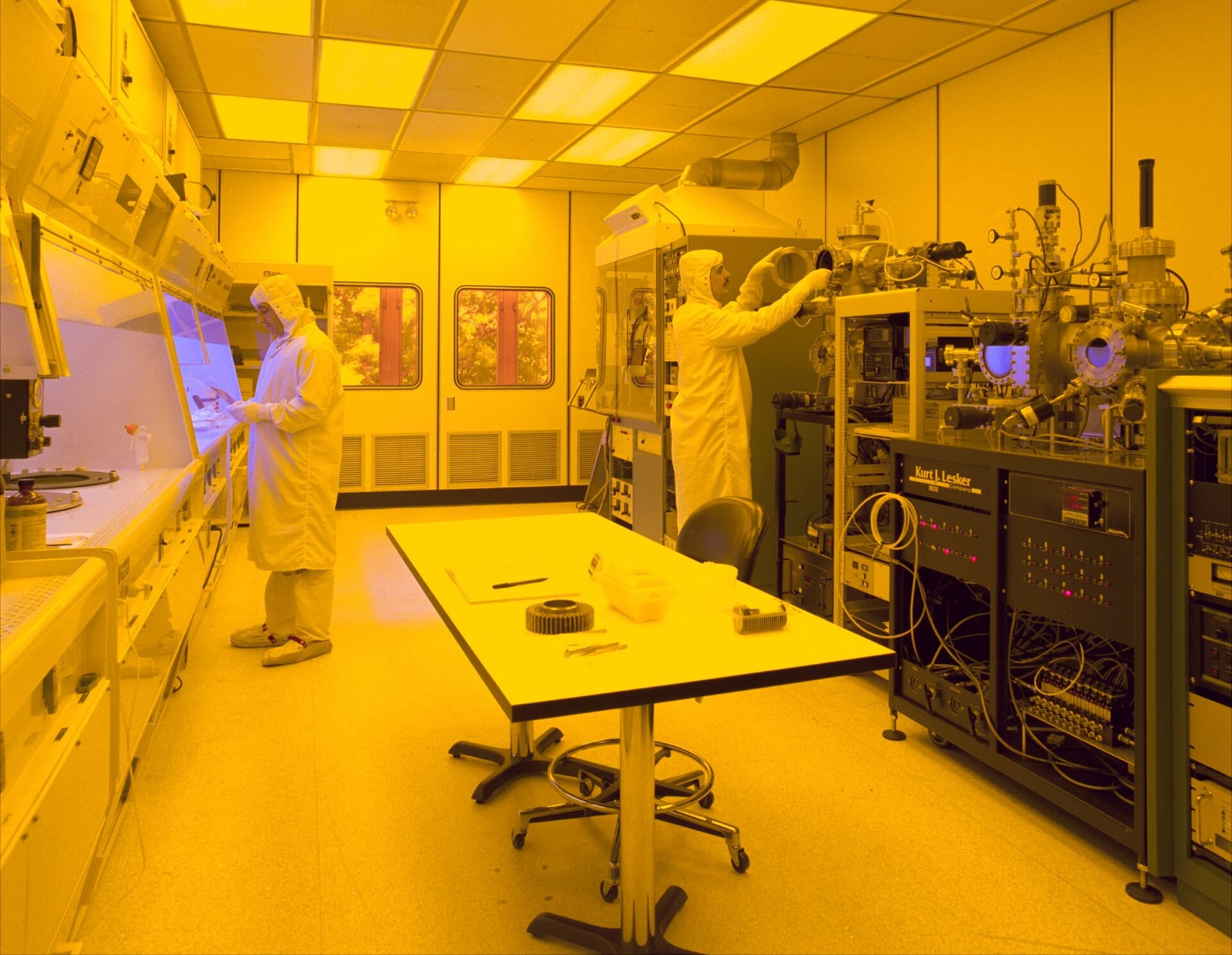 Two technicians in full cleanroom suits working under yellow lithography lighting in a semiconductor fabrication cleanroom