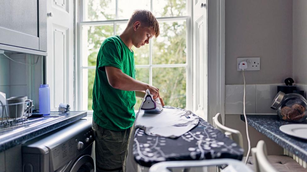 A person ironing beside a plug socket and washing machine in a kitchen