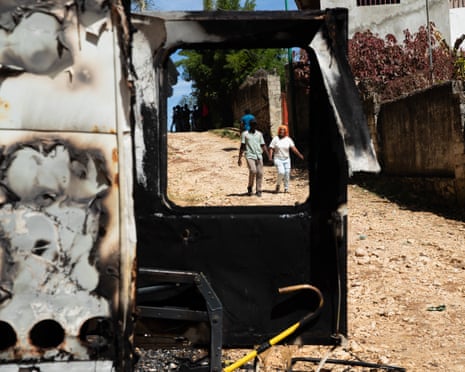People walking past a burnt-out vehicle in a street